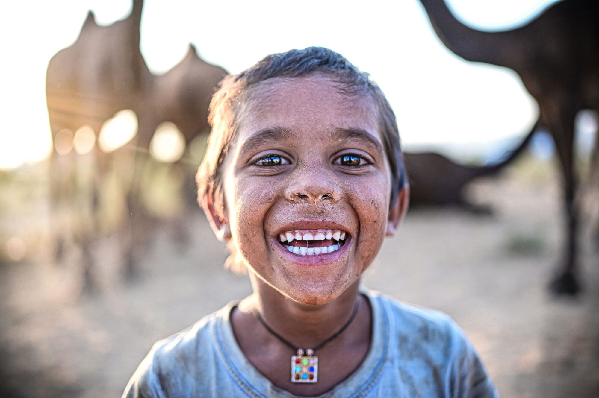 A cheerful child with a dirty face smiles widely in the Pushkar desert, surrounded by camels and warm light.