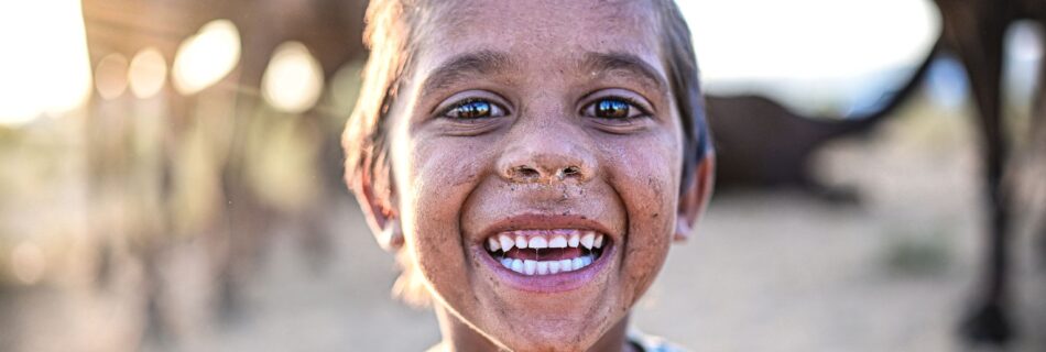 A cheerful child with a dirty face smiles widely in the Pushkar desert, surrounded by camels and warm light.