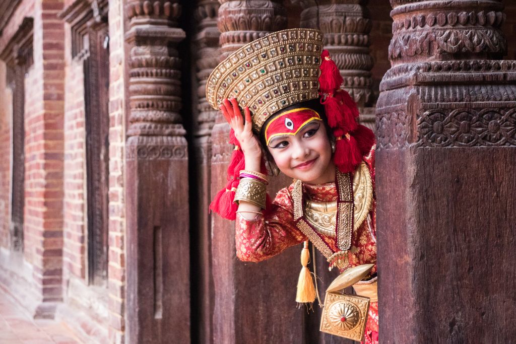 A young girl in traditional Nepalese dress peeks through ornate wooden architecture.