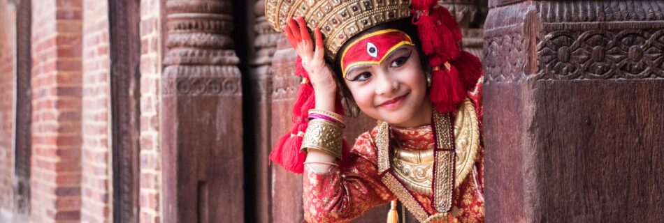 A young girl in traditional Nepalese dress peeks through ornate wooden architecture.