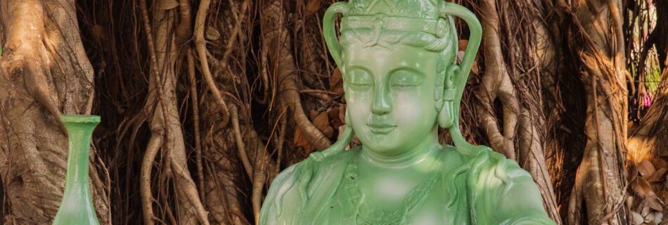 Green Buddha statue in a serene outdoor setting against a large tree.